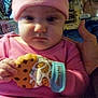 baby, child, pink_clothing, hat, teething_mitten, cookie_pattern, indoor, furniture, curtain, face, hand, person, clothing, expression, seated, room, background, household_items, portrait, serious