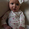 baby, child, onesie, headwear, bow, cherry_pattern, expression, face, hand, sitting, indoors, clothing, infant, cute, portrait, skin, person, young, wide_eyes, curious