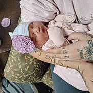 Nellie joined the competition — help win amazing prizes! newborn, baby, sleeping, hat, flower, tattoos, arms, cushion, blanket, person, indoor, soft_clothing, patterned_fabric, table, container, hand, resting, closeup, peaceful, infant