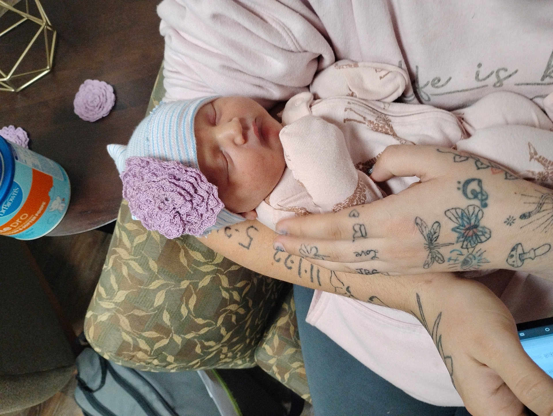 Nellie joined the competition — help win amazing prizes! newborn, baby, sleeping, hat, flower, tattoos, arms, cushion, blanket, person, indoor, soft_clothing, patterned_fabric, table, container, hand, resting, closeup, peaceful, infant