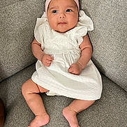 Léna a rejoint le concours — aidez-le/la à gagner de superbes lots ! baby, infant, child, white_dress, pink_headband, bow, couch, gray_couch, sitting, curious_expression, chubby_cheeks, indoors, soft_lighting, portrait, cute, small_hands, barefoot, comfortable, casual, young