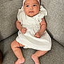 baby, infant, child, white_dress, pink_headband, bow, couch, gray_couch, sitting, curious_expression, chubby_cheeks, indoors, soft_lighting, portrait, cute, small_hands, barefoot, comfortable, casual, young
