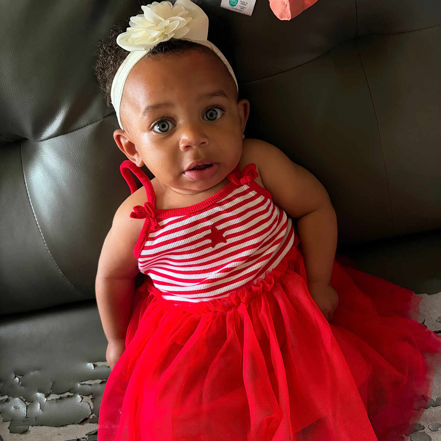 Laniya is registered to the contest to win money with this photo: toddler, child, red_dress, headband, flower, striped_clothing, sitting, leather_couch, curious_expression, indoor, person, fashion, cute, young_child, casual, portrait, face, eyes, hair, couch