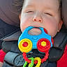 baby, child, car_seat, toy, teether, red_shirt, hair, face, seatbelt, infant, closeup, indoors, safety, child_seat, colorful, plastic_toy, cute, young_child, baby_toy, car_interior