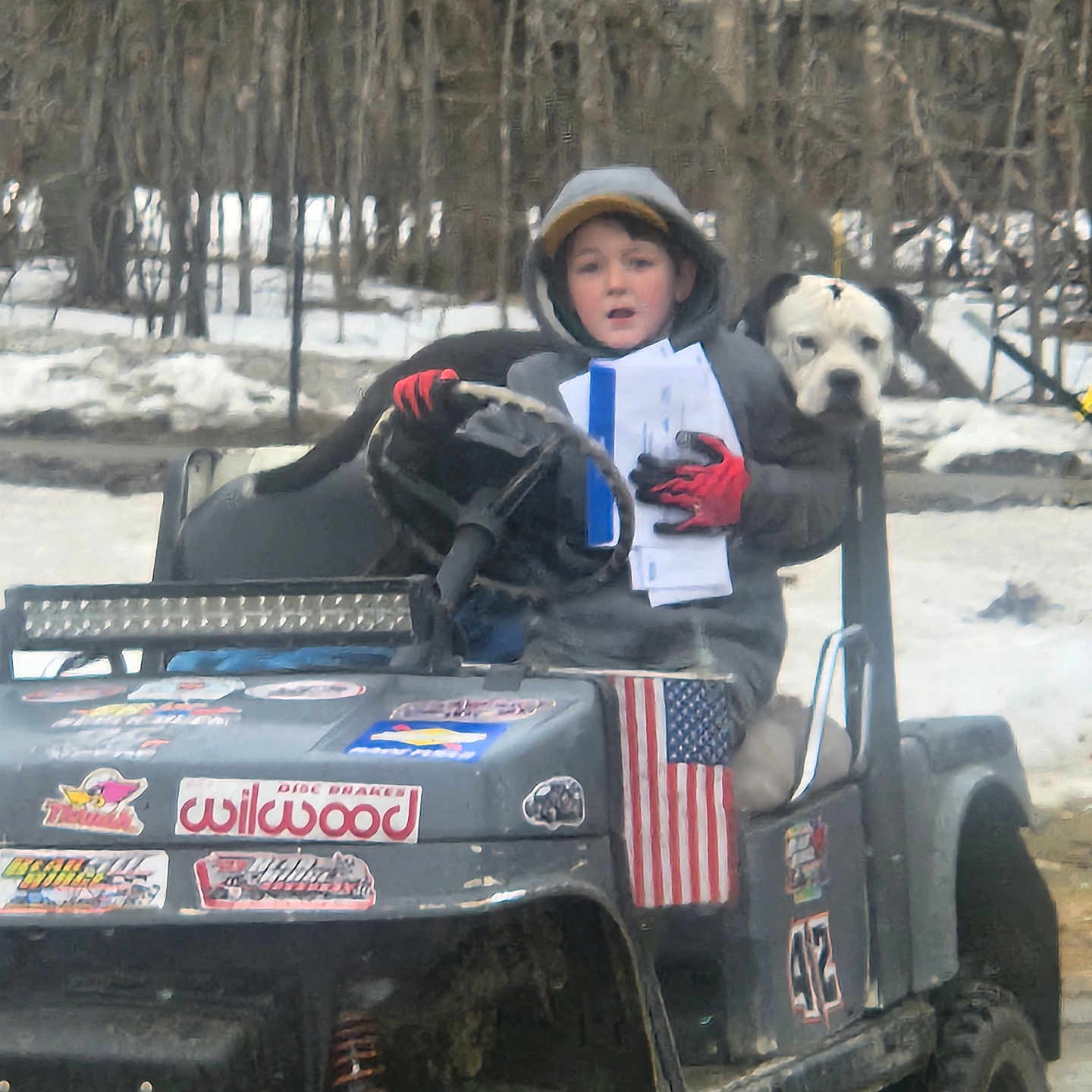 Brytton joined the competition — help win amazing prizes! child, dog, off_road_vehicle, hoodie, gloves, papers, stickers, american_flag, snow, forest, trees, outdoor, winter, vehicle, steering_wheel, pet, animal, person, glove, nature