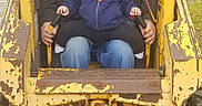 Brytton is registered to the contest to win money with this photo: child, adult, construction_vehicle, yellow, outdoor, hat, hoodie, dinosaur_hoodie, beard, overalls, rust, metal, controls, machine, person, safety, playful, family, toddler, elderly