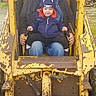 child, adult, construction_vehicle, yellow, outdoor, hat, hoodie, dinosaur_hoodie, beard, overalls, rust, metal, controls, machine, person, safety, playful, family, toddler, elderly