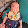 baby, infant, bib, onesie, pacifier, blanket, cushion, face, chubby_cheeks, blue_onesie, patterned_clothing, indoor, person, cute, portrait, child, sitting, skin, hand, expression