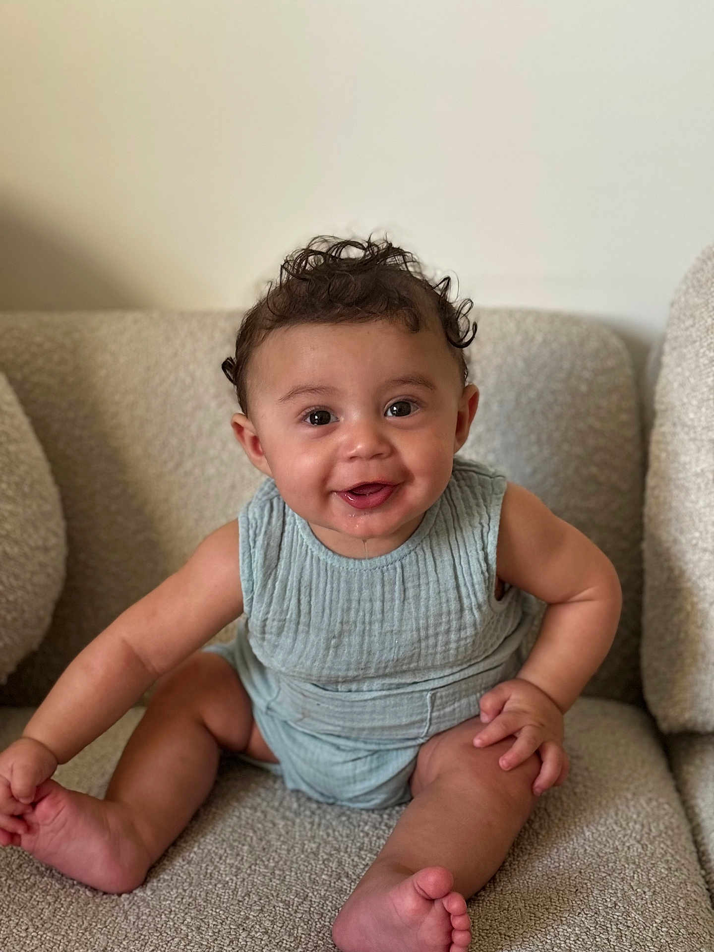 Elyah a rejoint le concours — aidez-le/la à gagner de superbes lots ! baby, child, smiling, curly_hair, sitting, couch, indoors, portrait, happy, cute, infant, clothing, skin, face, eyes, mouth, drool, furniture, light_blue, sleeveless