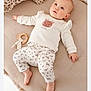 baby, infant, child, lying_down, cushion, toy, wooden_toy, clothing, white_clothes, patterned_pants, deer_design, soft_texture, indoors, cute, gazing_up, barefoot, pink_cheeks, comfortable, relaxed, nursery