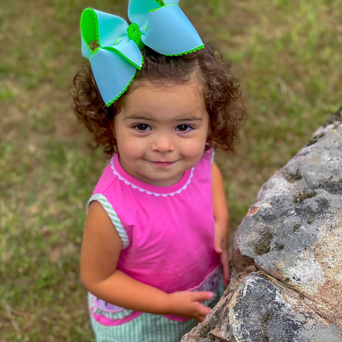 Edith joined the competition — help win amazing prizes! bow, casual, child, curly_hair, cute, daylight, girl, grass, green_clothing, happy, moss, nature, outdoor, person, pink_clothing, portrait, smile, standing, stone_wall, young_child