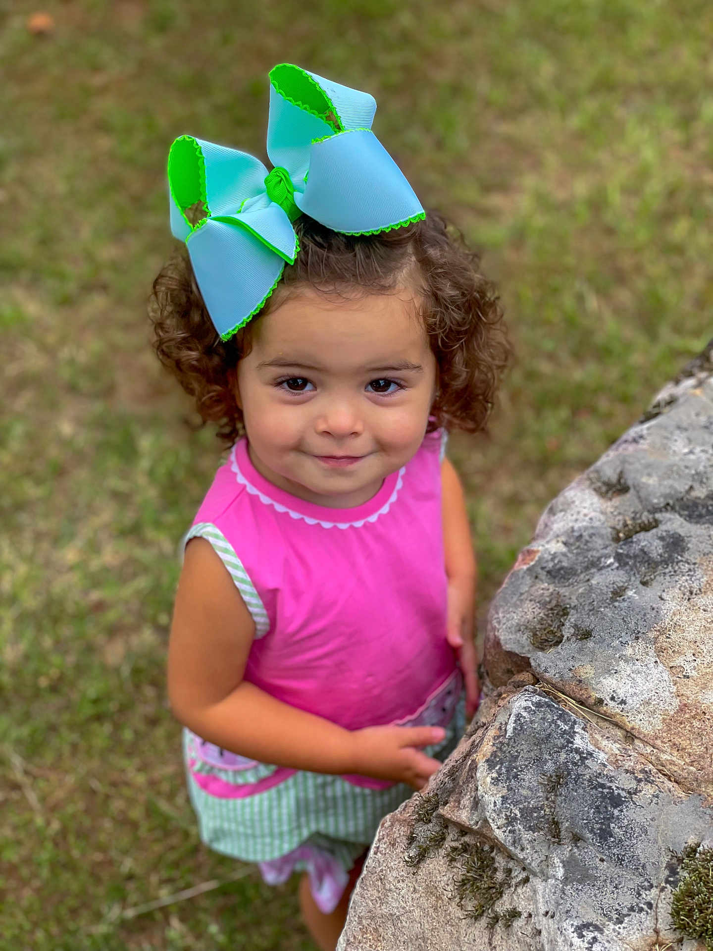 Edith joined the competition — help win amazing prizes! child, girl, curly_hair, bow, pink_clothing, green_clothing, outdoor, grass, stone_wall, moss, smile, portrait, cute, standing, person, young_child, nature, daylight, happy, casual