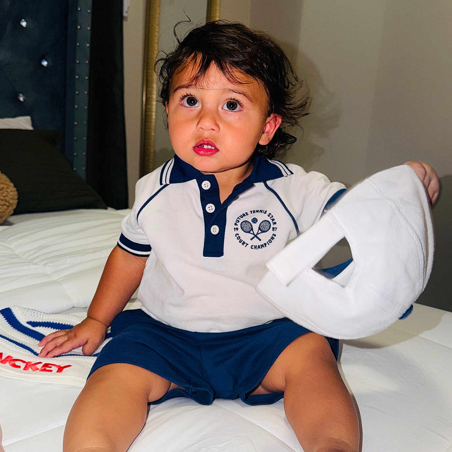 Bryant is registered to the contest to win money with this photo: toddler, child, bed, white_cap, shorts, polo_shirt, indoor, curly_hair, large_eyes, barefoot, blanket, mirror, bedroom, furniture, portrait, sitting, curious, clothing, hand, young_child