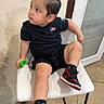 child, toddler, portrait, sitting, chair, black_shirt, black_shorts, sneakers, red_shoes, green_object, fashion, casual_clothing, indoors, wall, door, window, tile_floor, looking_away, single_person, full_body