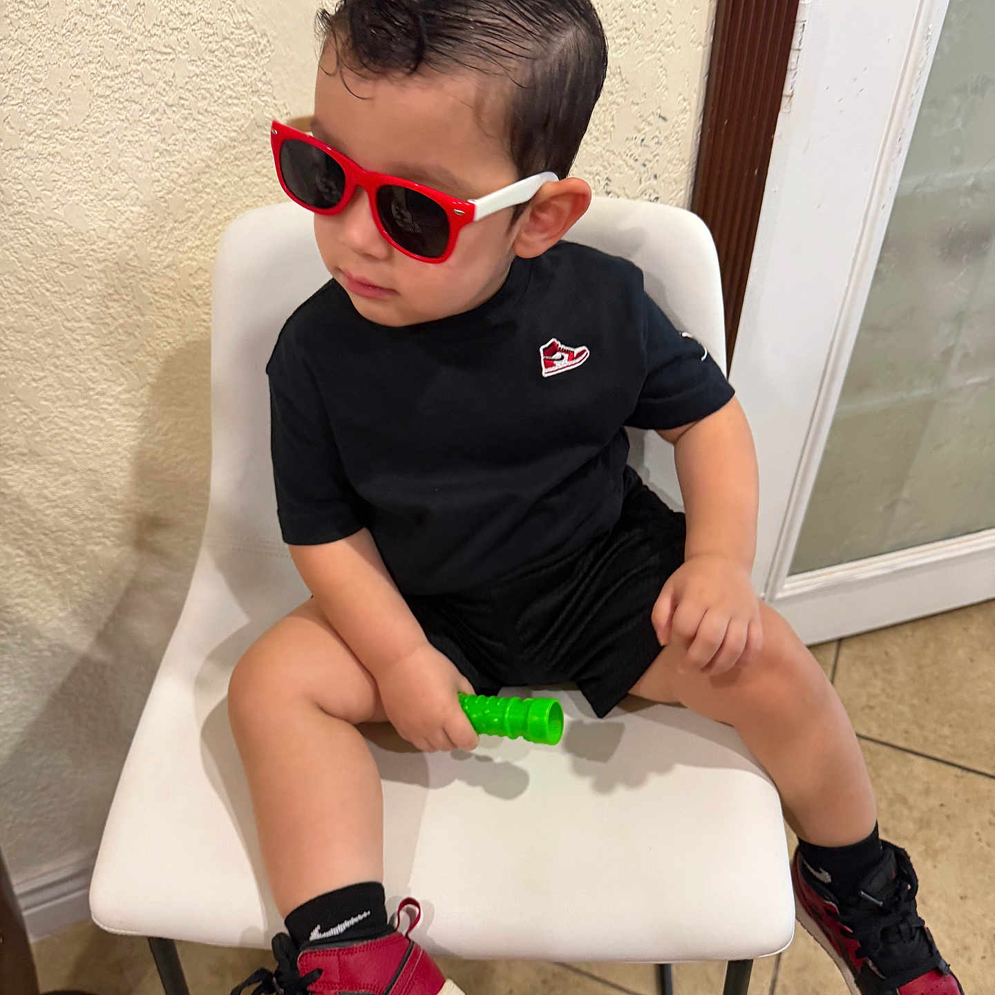 Anders Corria joined the competition — help win amazing prizes! child, toddler, sitting, sunglasses, red_sunglasses, black_clothing, red_shoes, sneakers, toy, green_toy, chair, indoor, portrait, fashion, stylish, short_hair, smiling, casual, tile_floor, door
