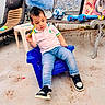 child, toddler, sitting, chair, blue_chair, pink_shirt, jeans, sneakers, toy, yellow_toy, soccer_ball, toy_blaster, outdoor, patio, bench, mural, furniture, portrait, serious_expression, fashion