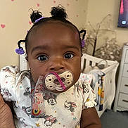 Naomi is registered to the contest to win money with this photo: baby, infant, pacifier, hair_ties, curly_hair, minnie_mouse, onesie, hand, nursery, crib, blanket, wall_decals, hearts, furniture, indoor, child, cute, close_up, person, portrait