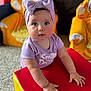 Amelia is registered to the contest to win money with this photo: baby, child, headband, bow, purple_clothing, red_cushion, yellow_cushion, plush_chair, carpet, indoor, furniture, person, cute, infant, hands, face, eyes, expression, soft_lighting, portrait