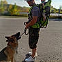 man, baby, dog, german_shepherd, sunglasses, cap, baby_carrier, leash, outdoor, road, sunny, shadow, green, camouflage_shorts, white_shoes, pacifier, headband, sky, trees, people