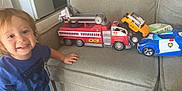 Dominic is registered to the contest to win money with this photo: toddler, child, toy, fire_truck, police_car, yellow_suv, couch, indoor, smiling, blue_shirt, shorts, play, vehicle, furniture, car_toy, happy, kid, living_room, person, casual