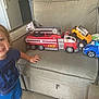 toddler, child, toy, fire_truck, police_car, yellow_suv, couch, indoor, smiling, blue_shirt, shorts, play, vehicle, furniture, car_toy, happy, kid, living_room, person, casual