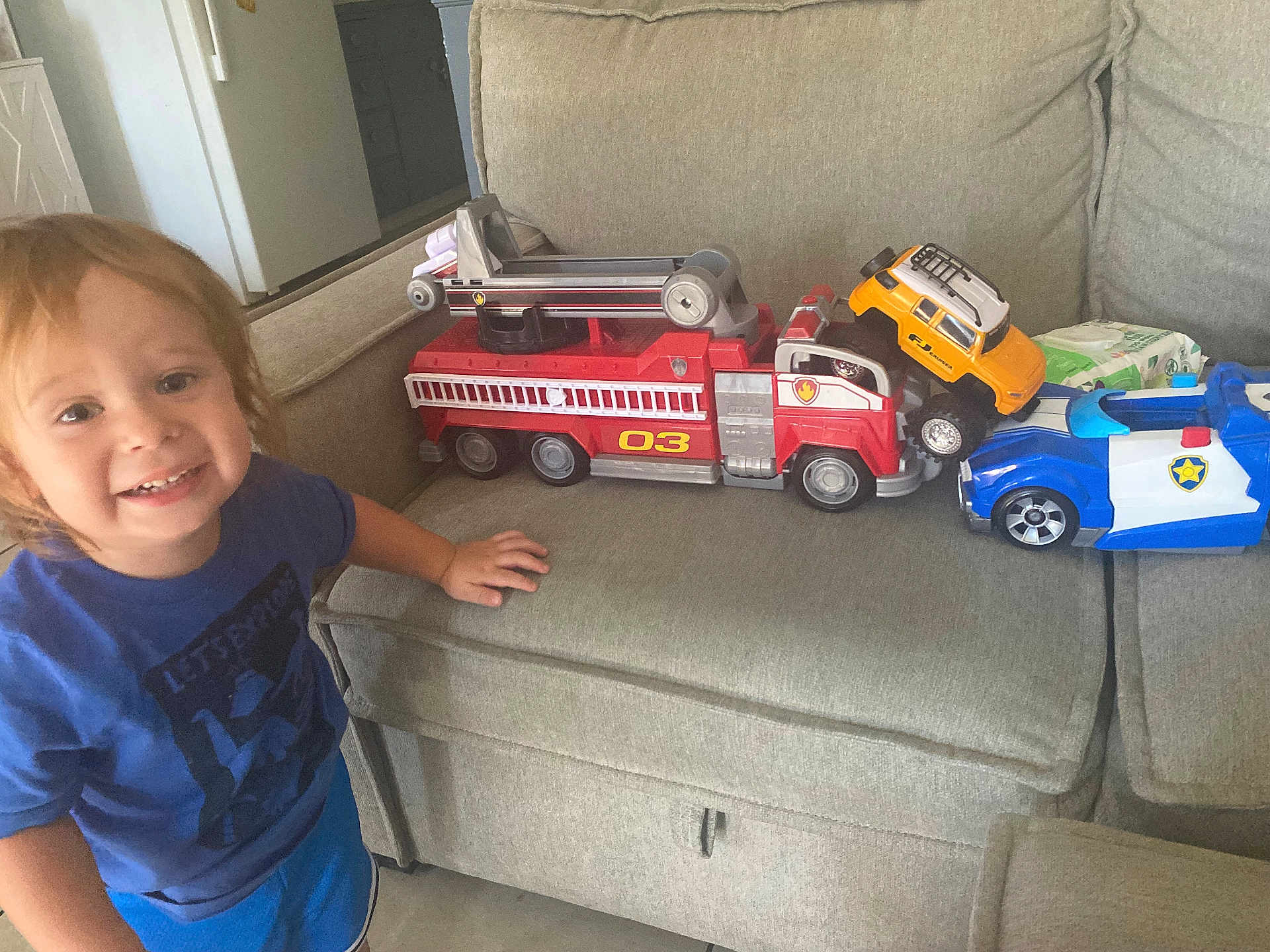 Dominic is registered to the contest to win money with this photo: toddler, child, toy, fire_truck, police_car, yellow_suv, couch, indoor, smiling, blue_shirt, shorts, play, vehicle, furniture, car_toy, happy, kid, living_room, person, casual