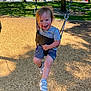 child, toddler, swing, playground, outdoor, sunny, wood_chips, tree, grass, smiling, happy, casual_clothing, shorts, tshirt, sneakers, metal_chain, daytime, park, person, recreation