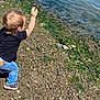 Dominic is registered to the contest to win money with this photo: toddler, child, river, water, rocks, seaweed, shoreline, outdoor, nature, casual_clothing, jeans, black_shirt, sneakers, throwing, playing, sunny, daylight, young_child, side_view, activity