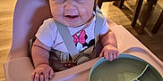 Jade is registered to the contest to win money with this photo: baby, high_chair, smiling, indoor, plate, divided_plate, mickey_mouse, clothing, person, child, wood_floor, table, chair, dining_room, happy, cute, feeding, meal, infant, straps