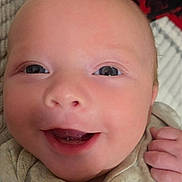 John is registered to the contest to win money with this photo: baby, smiling, close_up, face, infant, eyes, hand, fist, clothing, blanket, soft, cute, portrait, skin, head, happy, newborn, young, person, indoors