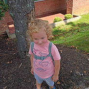 Myles is registered to the contest to win money with this photo: child, toddler, curly_hair, pink_shirt, dinosaur_print, shorts, blue_shoes, backpack_straps, tree, mulch, grass, brick_building, sidewalk, outdoor, daylight, smile, standing, nature, shy, person