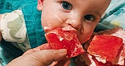 Frank is registered to the contest to win money with this photo: adult_hand, baby, bed, blanket, blue_clothing, child, curious, eating, eyes, face, family, fruit, green_toy, hand, indoors, infant, portrait, red_fruit, snack, watermelon