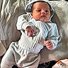 baby, newborn, child, portrait, lying_down, white_clothing, hood, onesie, blanket, pillow, bed, indoor, sunlight, face, eyes_open, serious_expression, hands, infant, soft_fabric, cozy