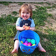 Ricky is registered to the contest to win money with this photo: toddler, child, grass, bucket, plastic_eggs, outdoor, play, happy, tongue_out, blue_bucket, colorful, summer, casual, nature, smile, person, barefoot, denim_overalls, short_sleeve, fun