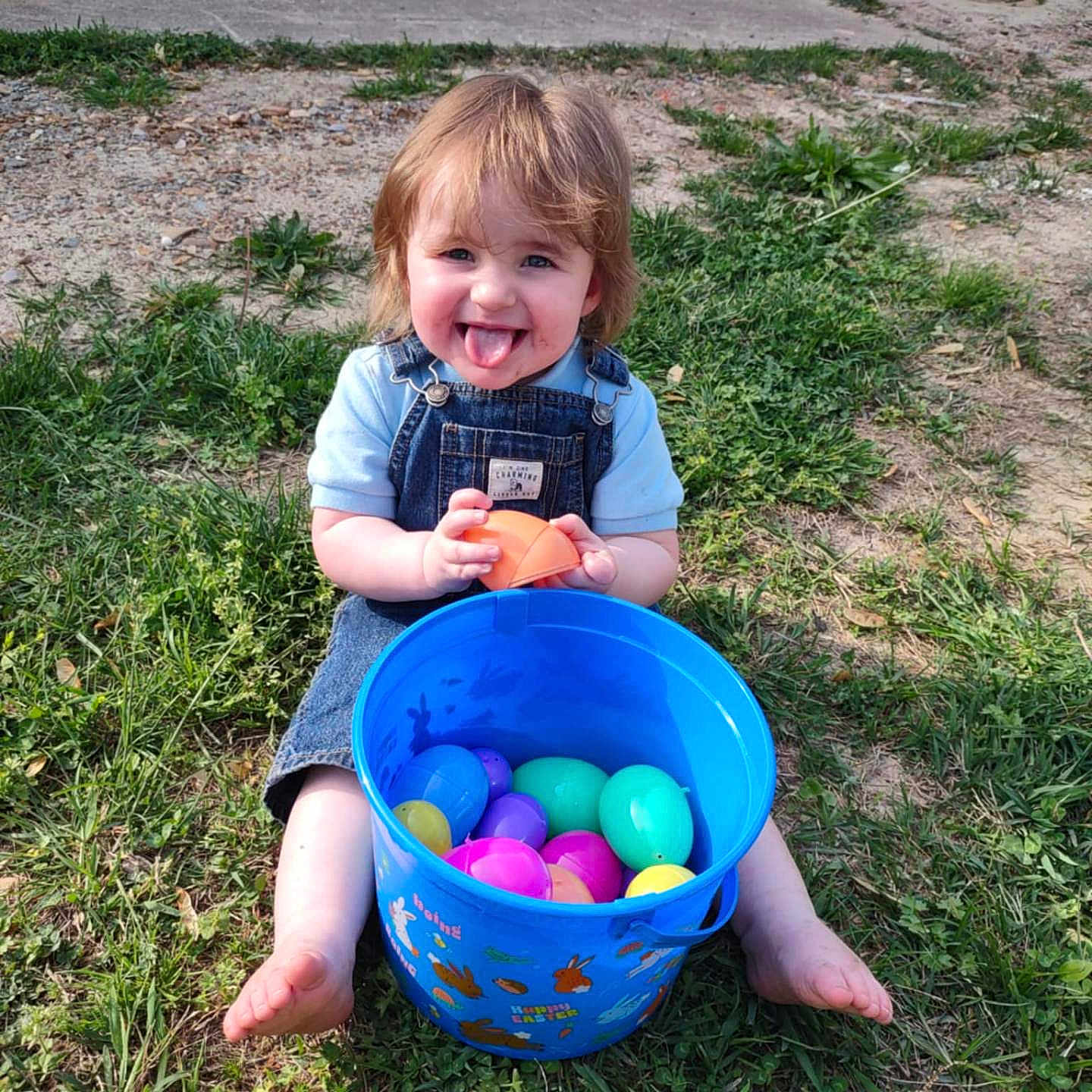 Ricky is registered to the contest to win money with this photo: toddler, child, grass, bucket, plastic_eggs, outdoor, play, happy, tongue_out, blue_bucket, colorful, summer, casual, nature, smile, person, barefoot, denim_overalls, short_sleeve, fun