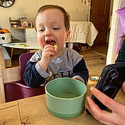 Alex a rejoint le concours — aidez-le/la à gagner de superbes lots ! child, toddler, eating, snack, green_bowl, wooden_table, phone, hand, high_chair, living_room, wall_clock, wooden_door, smile, candid, mealtime, furniture, cup, clothing, portrait, messy_room