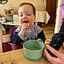 child, toddler, eating, snack, green_bowl, wooden_table, phone, hand, high_chair, living_room, wall_clock, wooden_door, smile, candid, mealtime, furniture, cup, clothing, portrait, messy_room