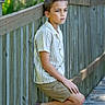 boy, brown_shoes, casual_clothing, child, daylight, expression, fence, footwear, greenery, leaning, nature, outdoor, portrait, shirt, shorts, side_view, summer, thoughtful, wood, young