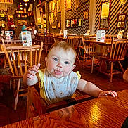 Shiloh joined the competition — help win amazing prizes! baby, child, high_chair, restaurant, wooden_chair, wooden_table, bib, playful, tongue_out, blue_eyes, indoor, people, furniture, decor, warm_lighting, portrait, smiling, casual_clothing, family, dining