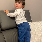 Mathys participe au concours pour gagner de l'argent avec cette photo : toddler, child, couch, jeans, white_shirt, socks, pillow, indoor, curly_hair, standing, leather_furniture, beige_wall, holding, looking_back, casual_clothing, home, young_child, small_person, soft_lighting, cute
