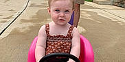 Claire joined the competition — help win amazing prizes! toddler, child, toy_car, pink, steering_wheel, adult, hands, yellow_shirt, shorts, driveway, concrete, outdoor, greenery, summer, playtime, person, vehicle, expression, casual, daylight