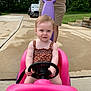 toddler, child, toy_car, pink, steering_wheel, adult, hands, yellow_shirt, shorts, driveway, concrete, outdoor, greenery, summer, playtime, person, vehicle, expression, casual, daylight