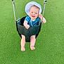 Claire is registered to the contest to win money with this photo: baby, child, swing, playground, green_grass, smile, happy, hat, sun_hat, outdoor, infant, legs, barefoot, blue_clothing, metal_chains, fun, joy, seat, play, park