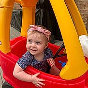 Claire is registered to the contest to win money with this photo: toddler, child, toy_car, red, yellow, headband, pink, smile, outdoor, deck, play, cute, happy, person, plastic, small_wheels, blue_button, short_sleeve, patterned_shirt, curly_hair