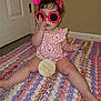 baby, child, indoor, sitting, pink, headband, sunglasses, crocheted_blanket, milestone_plaque, nine_months, curly_hair, floor, carpet, person, cute, playful, clothing, hand, legs, door