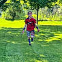 child, face_paint, red_shirt, shorts, grass, tree, sunlight, running, outdoor, patriotic, flag_pattern, gloves, cape, socks, smile, boy, summer, recreation, fun, nature