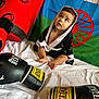 baby, barefoot, black, blue, boxing_bag, boxing_gloves, child, costume, curious, flag, footwear, green, hooded_robes, indoors, playful, red, red_circle, sitting, white_sheet, young_child