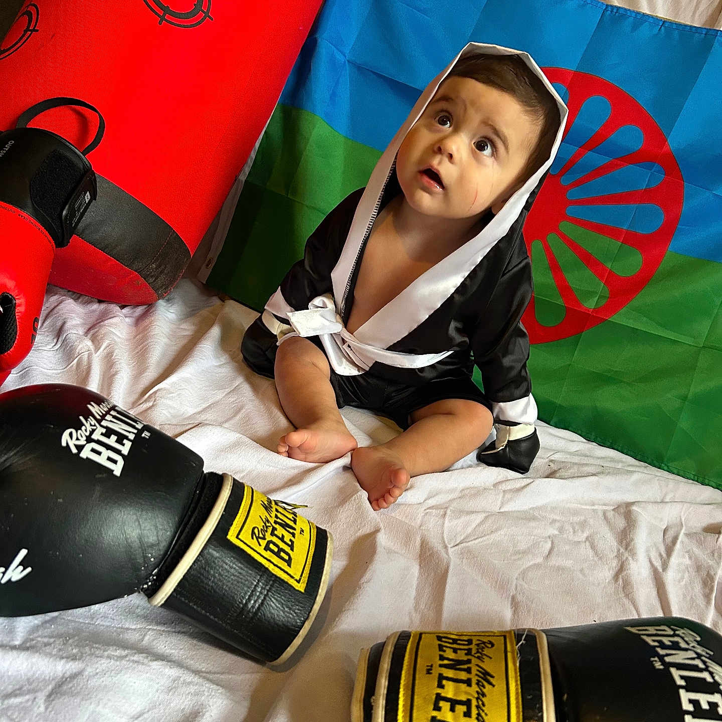 Keyson participe au concours pour gagner de l'argent avec cette photo : baby, barefoot, black, blue, boxing_bag, boxing_gloves, child, costume, curious, flag, footwear, green, hooded_robes, indoors, playful, red, red_circle, sitting, white_sheet, young_child