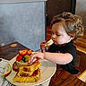 Heavenly joined the competition — help win amazing prizes! child, toddler, portrait, eating, sandwich, bread, cheese, fruit, fruit_bowl, strawberry, grapes, table, plate, indoor, restaurant, food, sitting, curly_hair, black_shirt, focused