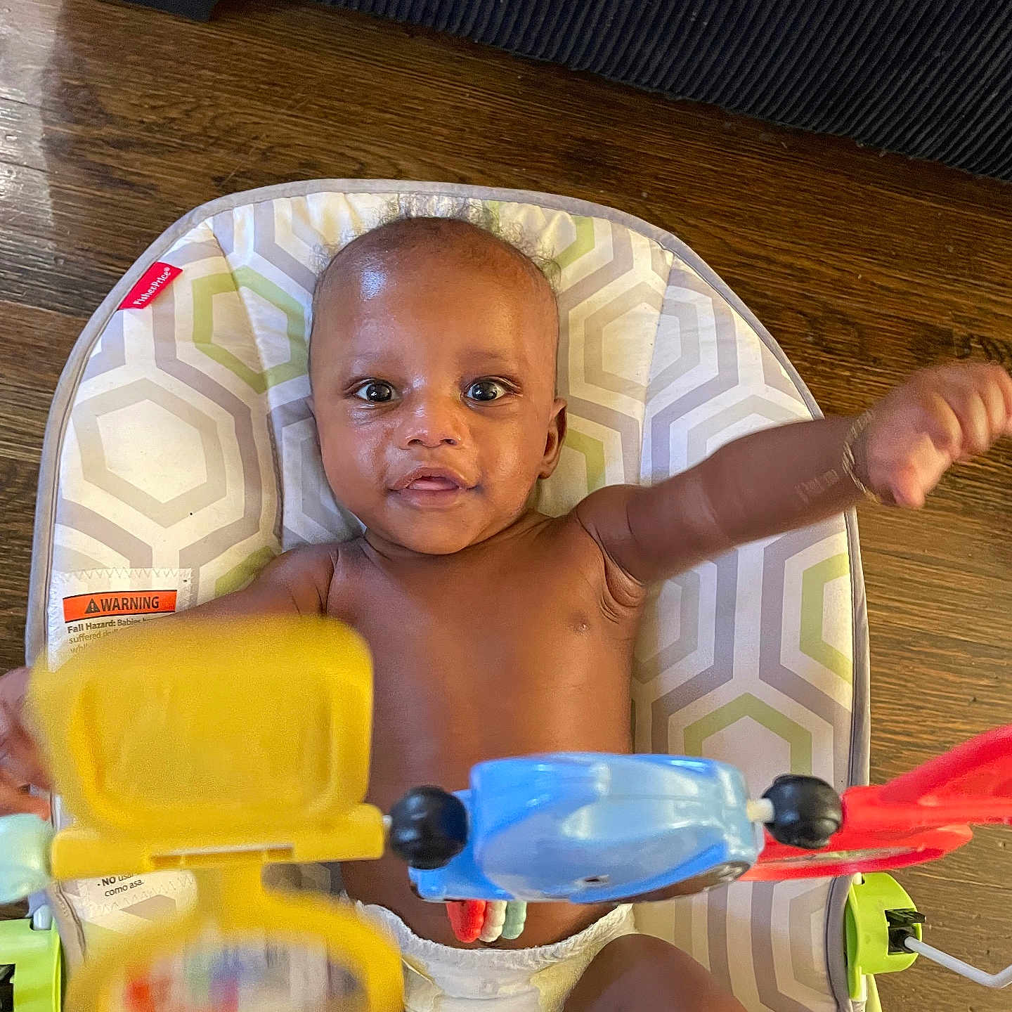 Marvis is registered to the contest to win money with this photo: arm, baby, baby_seat, child, colorful, cute, diaper, eyes, face, floor, furniture, hand, indoor, infant, pattern, person, play, smile, toy, wood_floor